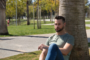 Obraz premium Young and handsome man, with blue eyes, beard, green t-shirt and jeans, sitting and leaning on a palm tree trunk and with a distracted gesture. Concept beauty, fashion, trend, thoughts, distraction.