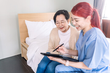 Obraz premium Asian woman nurse wearing scrubs report the health status of Senior Asian woman with digital tablet in the bedroom. Caregiver visit at home. Home health care and nursing home concept.