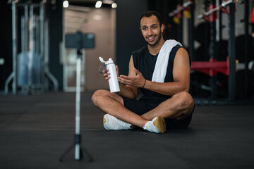 Fototapeta na wymiar Sport Accessories. Black Male Fitness Blogger Showing Water Bottle At Smartphone Camera