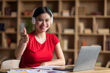 Glad young asian woman with down syndrome in headphones with laptop point finger at camera in office