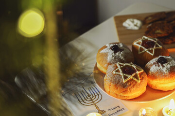 Close-up selective focus on a sufganiya donut with patterns in the form of a star of David made of powdered sugar, placed on a plate for the holiday of Hanukkah next to candles.
