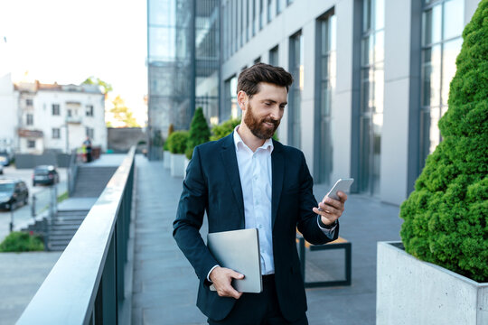 Smiling confident young businessman with beard in suit with laptop typing on smartphone