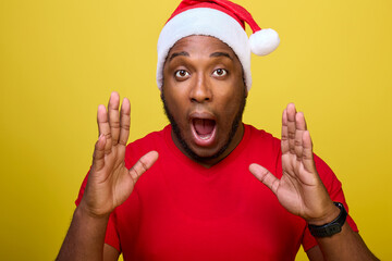Shocked young African-American Santa Claus in a red T-shirt and a Christmas hat, shouting with a hand gesture at his mouth, isolated on a yellow background. Concept Happy New Year, Merry Christmas