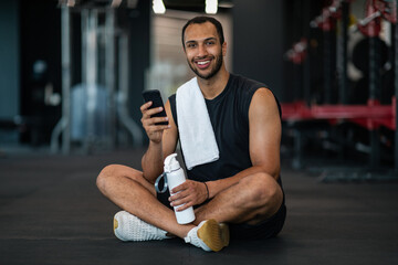 Fototapeta na wymiar Smiling Black Male Athlete Relaxing With Smartphone After Training In Gym