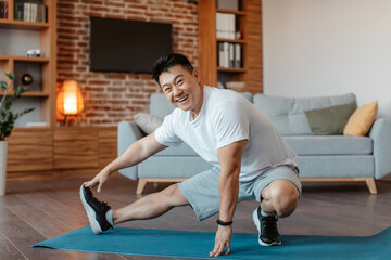 Fototapeta premium Happy mature sportsman making exercises for legs, stretching on fitness mat in living room and smiling at camera