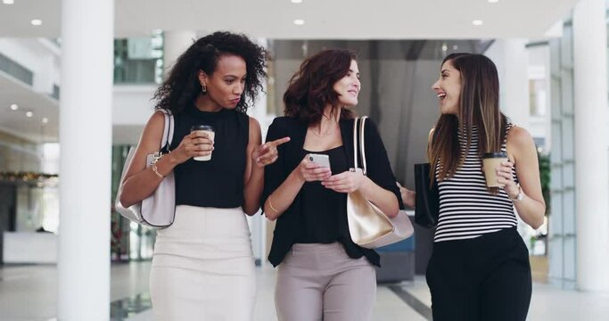 Female business women talking, chatting and checking social media on phone while walking inside an office. Laughing, smiling and cheerful colleagues gossiping and enjoying their coffee break together