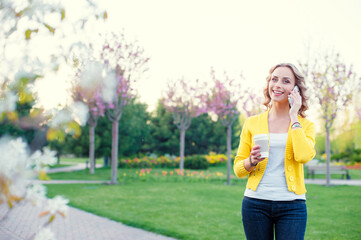 Walking by spring park. Attractive young woman talking by phone and holding cup of coffee outdoors.