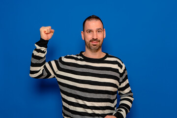 Young handsome man wearing striped sweater standing over isolated blue background angry and mad raising fist frustrated. Rage and aggressive concept