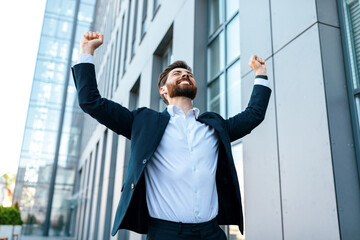 Cheerful attractive young caucasian businessman with beard in suit raises his hands up and rejoices...