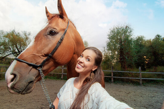 Funny selfie with my friend! Attractive smiling young woman holding camera and making selfie with her horse outdoors.