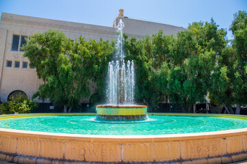 a tall flowing water fountain with blue water surrounded by lush green trees with blue sky in Pasadena California USA