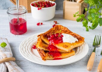 Finnish pancake pannukakku with redcurrant jam and sour cream or whipped cream on a white ceramic plate on a light wooden background. Scandinavian cuisine