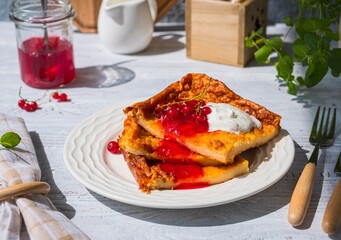 Finnish pancake pannukakku with redcurrant jam and sour cream or whipped cream on a white ceramic plate on a light wooden background. Scandinavian cuisine