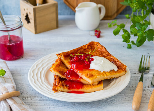 Finnish pancake pannukakku with redcurrant jam and sour cream or whipped cream on a white ceramic plate on a light wooden background. Scandinavian cuisine