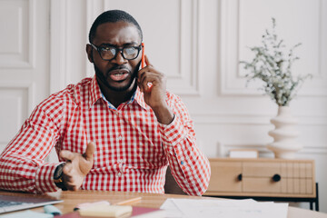 Dissatisfied African American businessman emotionally speaking on smartphone while sitting at desk