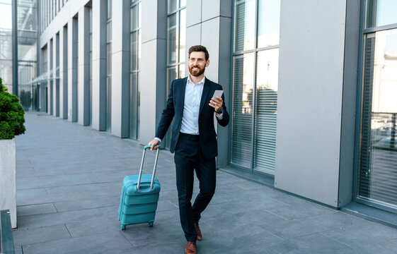 Smiling millennial european businessman with beard in suit and smartphone goes to airport with suitcase