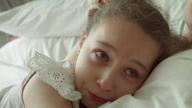 Portrait cute upset child little girl with big tears flowing down his cheeks,child lies at home on bed next to his mother and cries, tears flow with a screaming face.Mom takes pity on her daughter.