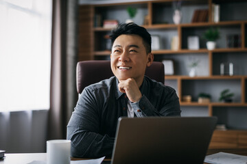 Smiling pensive adult asian businessman inspired by idea at laptop to workplace in home office...