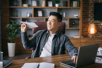 Cheerful handsome middle aged asian businessman play with paper plane at workplace at laptop in home