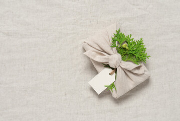 Christmas gift wrapped in fabric with thuja leaves and blank label, beige linen textile background. A traditional Japanese furoshiki gift. Zero waste concept. Top view, selective focus.