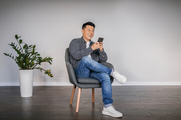 Happy adult asian man typing on smartphone sitting in armchair in minimalist living room interior,...