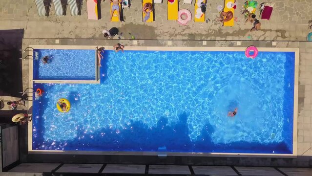 Aerial view from flying drone of people having fun, playing and resting in water in pool. kurt or villa with vacationers. Party of friends or celebration. Sun beds and young people sunbathe in sun.