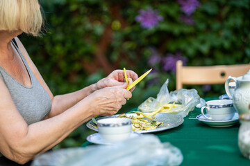 old woman peels green beans in the garden at the table - obrazy, fototapety, plakaty
