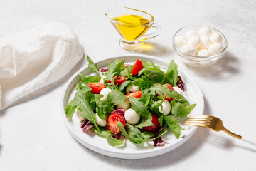 Fresh green salad made with mozzarella cheese, cherry tomatoes, arugula and olive oil on white stone table background. Healthy food and diet concept. Flat lay, top view.