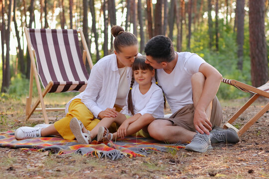 Image of young family enjoying moment while sitting on blanket in park and kissing their cute daughter, people relaxing in wood. spending weekend together.