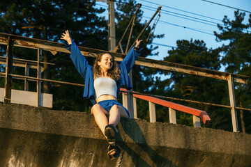 happy woman tourist in blue casual clothes and summer sandals sits at the top of the railway in the evening at sunset, she raised her hands up and rejoices