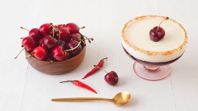 Dark and white chocolate mousse or panna cotta with chili cherry jelly in a glass with wooden bowl of ripe cherries on white wooden table. High angle view, no people.