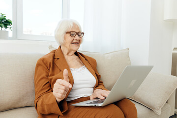 a happy, emotional elderly woman with gray hair is sitting in a bright apartment on a beige cozy sofa near the window happily working at a laptop showing a thumbs up