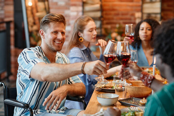 Portrait of smiling young man with disability clinking glasses with friends during dinner party