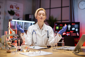 Fototapeta na wymiar Yong female scientist relaxing practicing yoga wearing lab coat working in laboratory while examining biochemistry sample in test tube and scientific instruments.