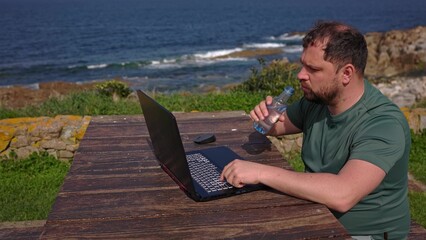 A man drinks a cart while working at a computer in nature.