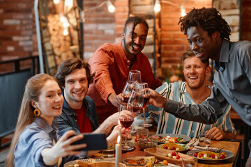 Diverse group of friends taking selfie photo at table during dinner party and clinking glasses
