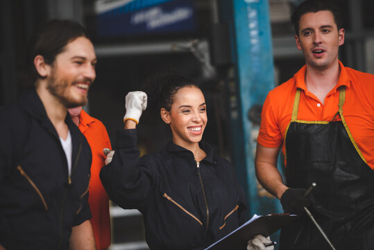 Group of young and senior male and female car mechanics in garage wearing uniform enjoying and celebrating with female worker holding clipping board