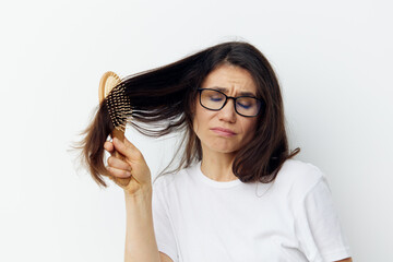 a sad, upset, depressed woman in a white T-shirt stands against a light background with glasses and tries to comb her long dark hair holding her palm on her face