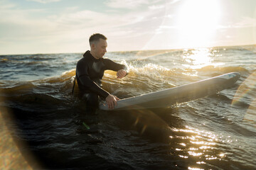 A male surfer in a wetsuit uses a sports watch on the waves at sunset in the sea