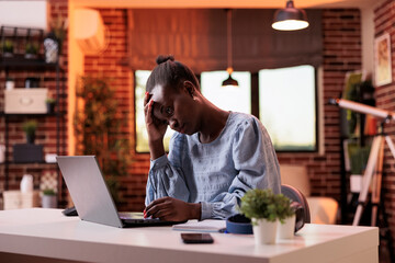 Overworked young female freelancer working on laptop in modern home office, burnout and workload concept. Stressed and exhausted african american businesswoman having headache