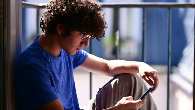 Footage of a handsome Caucasian young man sitting at the window using his mobile phone. Blurred background. Concept of leisure and use of technology by young people.
