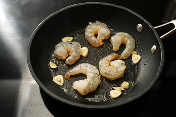 Raw peeled shrimps and garlic are sauteed in a cooking pan on the black stove, preparation for a delicious appetizer, copy space, selected focus