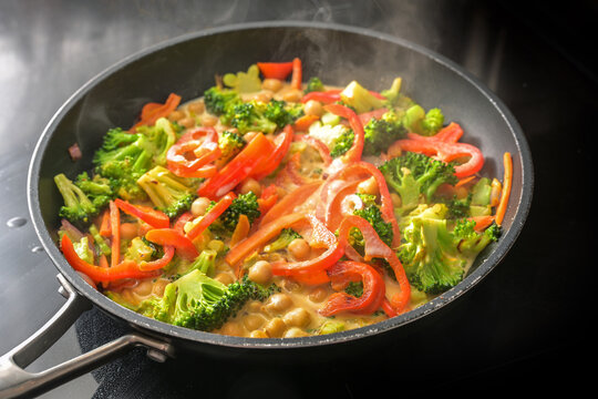Steaming vegetable curry with chickpeas, broccoli and red bell pepper in coconut milk in a cooking pan on a black stove, healthy meals for a vegan low carb diet, copy space, selected focus