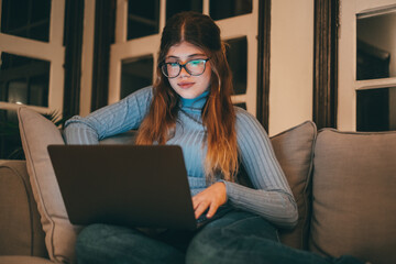 Pretty and beautiful young woman using laptop and surfing on the net at late night on the sofa at home. Attractive caucasian teenager girl with computer online. Lady wearing glasses working.