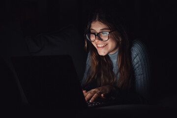 One happy cheerful young woman using laptop late at night in the living room on the sofa at home chatting. Online girl surfing the net revising social media. Smiling teenager using technology 