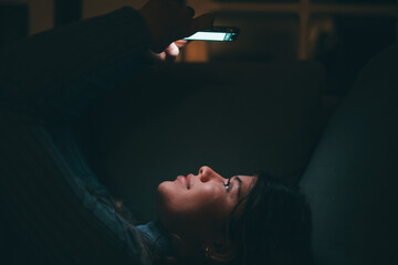 One beautiful young woman holding and using phone at home at late night on the sofa. Female teenager chatting with friends surfing the net online. Enjoying technology and internet