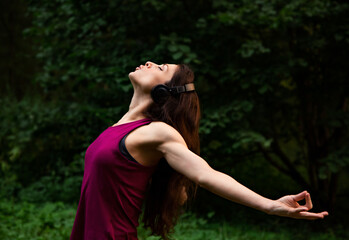 Sporty beautiful woman training yoga with lotus mudra outdoors summer green forest background and listening the music in headphone with relaxing closed calm eyes. Closeup