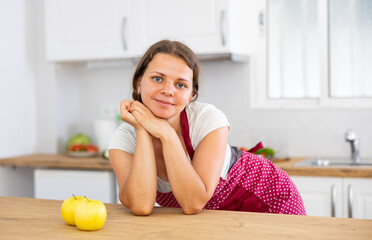 Fototapeta na wymiar Portrait of positive woman wearing apron, standing at kitchen table at home and looking at camera.