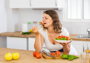 Obraz na płótnie Canvas Positive young woman eating freshly made vegetarian salad at kitchen countertop