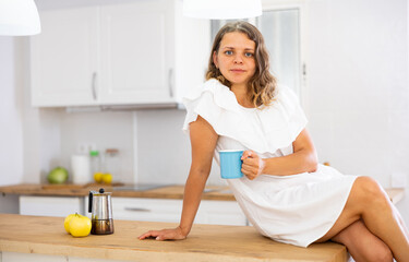Naklejka na ściany i meble Portrait of happy young woman in white dress drinking morning coffee in modern kitchen